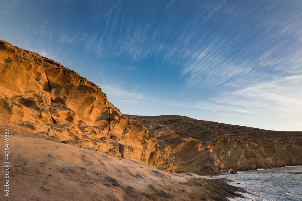 Brightly lit by the rising sun, a cliff of yellow sandstone on the seashore with large cracks and smooth lines of erosion. Thin threads of cirrus clouds in the blue sky. No people