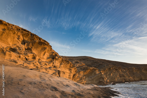 Brightly lit by the rising sun, a cliff of yellow sandstone on the seashore with large cracks and smooth lines of erosion. Thin threads of cirrus clouds in the blue sky. No people © Igor
