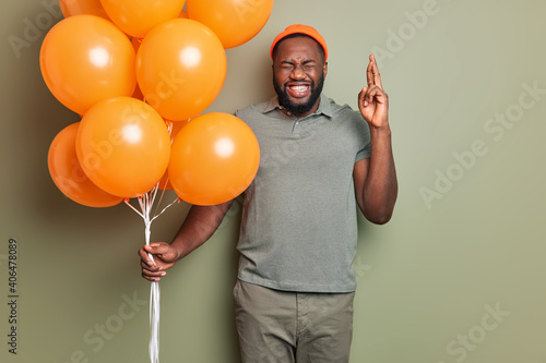 Happy bearded man with dark skin and beard crosses fingers makes wish on birthday holds bunch of bright orange inflated balloons dressed in stylish clothing stands indoor. People partying concept © wayhome.studio 