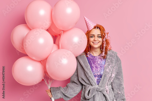 Cheerful redhead woman wears party hat and soft dressing gown has spoiled makeup holds inflated balloons spends free time on birthday party isolated over pink background accepts congratulations
