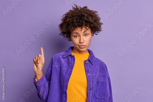 Serious African American woman has curly hair assures you to go upstairs indicates with index finger above wears fashionable clothes isolated over purple background. Look at this advertisement