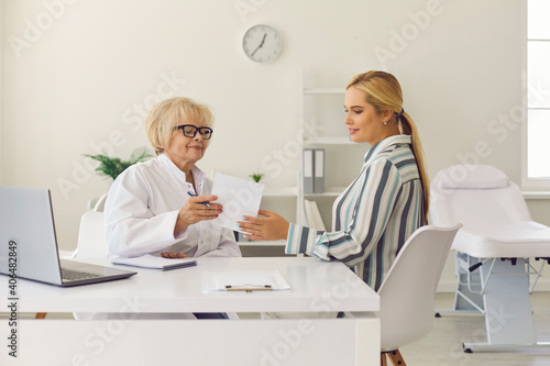 Positive confident senior woman doctor in white uniform giving medical prescriprion to young woman patient during consultation in medical clinic. Healthcare, communication with doctor concept