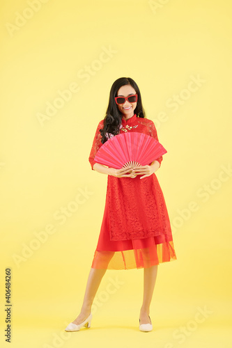 Smiling pretty young woman in red dress and sunglasses posing with fan, isolated on yellow