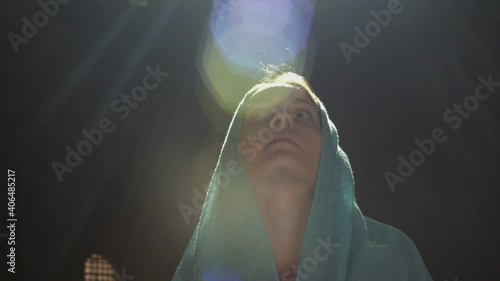 Young millennial woman in pink dress and blue headscarf stands in church, temple and prays, looks at the camera with a serious expression. Slow motion.