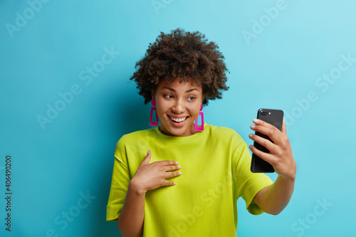 Pretty young African American woman holds modern mobile phone in front of herself has online conference makes video call connected to wireless internet enjoys positive talk wears green t shirt