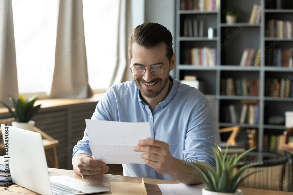 Smiling young businessman in eyeglasses looking at paper correspondence, reading pleasant good news. Excited happy male entrepreneur getting bank loan approvement notification at home office. Smiling young businessman in eyeglasses looking at paper correspondence, reading pleasant good news. Excited happy male entrepreneur getting bank loan approvement notification at home office.