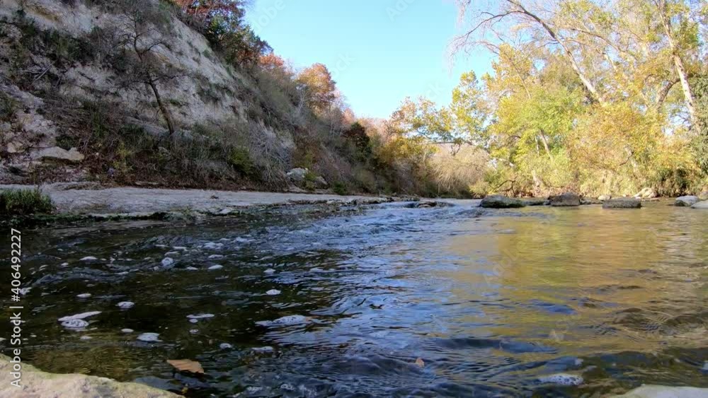 Panning left across a flowing creek. Rightmost trees and water are lit colorfully by the sun. A tall shaded cliff wall is on the left with exposed rock and vegetation. Panning left across a flowing creek. Rightmost trees and water are lit colorfully by the sun. A tall shaded cliff wall is on the left with exposed rock and vegetation.