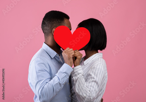 Side view of passionate black lovers kissing behind red paper heart, expressing their love on pink studio background