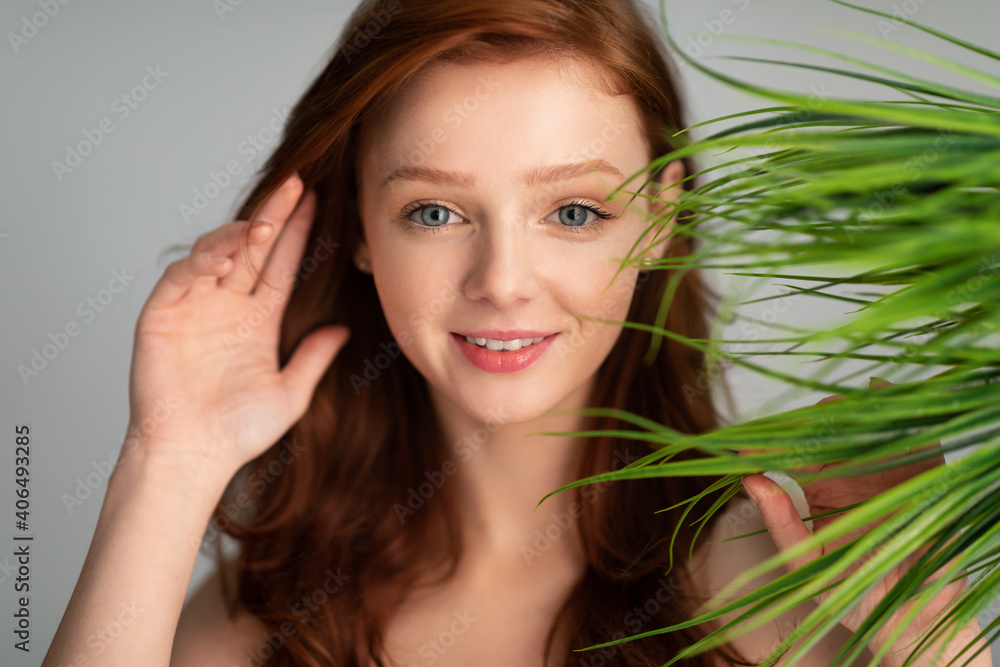 Fototapeta premium Ginger-Haired Young Woman Posing With Green Plant Leaves, Gray Background