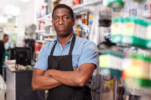 Portrait of confident experienced salesman in apron standing near racks with goods in household store