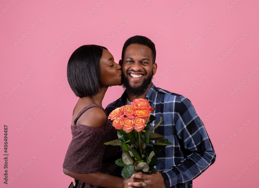 Affectionate young black woman with bouquet of flowers kissing her boyfriend on pink background. Valentine's Day Affectionate young black woman with bouquet of flowers kissing her boyfriend on pink background. Valentine's Day