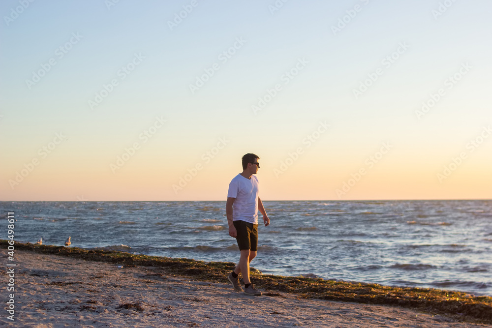 A young men in a white shirt and brown shorts walks along an empty beach at sunrise. A male in sunglases going on the shores of a calm morning sea and looking at sunrise.