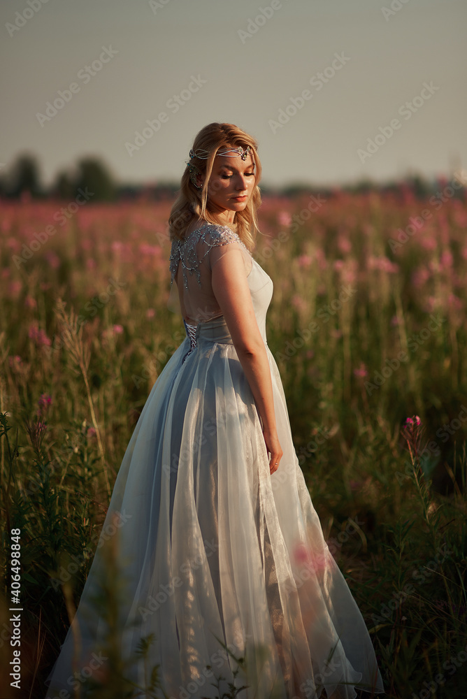 Romantic blonde woman running in a wonderful flower field. Warm sunset colors. Blue dress. Soft colors.