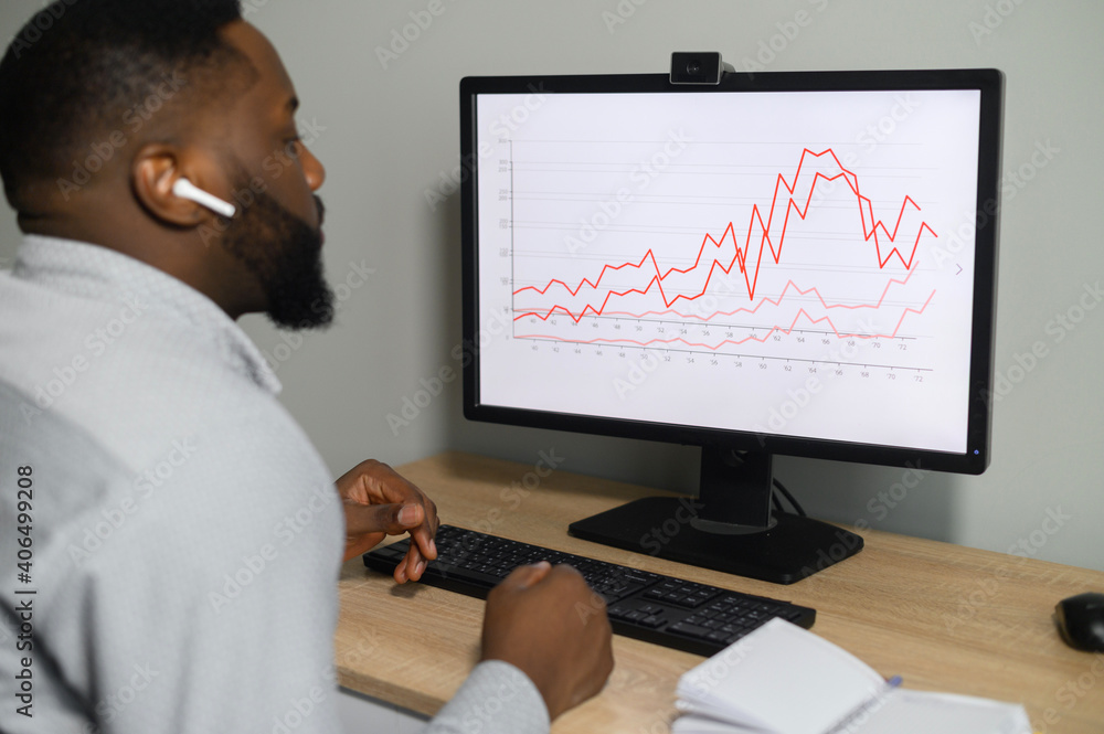 Concentrated African man in formal shirt works with a PC at office, a businessman analyzes the sales growth graph, verifies the datas. A multiracial male entrepreneur using PC at the desk, back view Concentrated African man in formal shirt works with a PC at office, a businessman analyzes the sales growth graph, verifies the datas. A multiracial male entrepreneur using PC at the desk, back view
