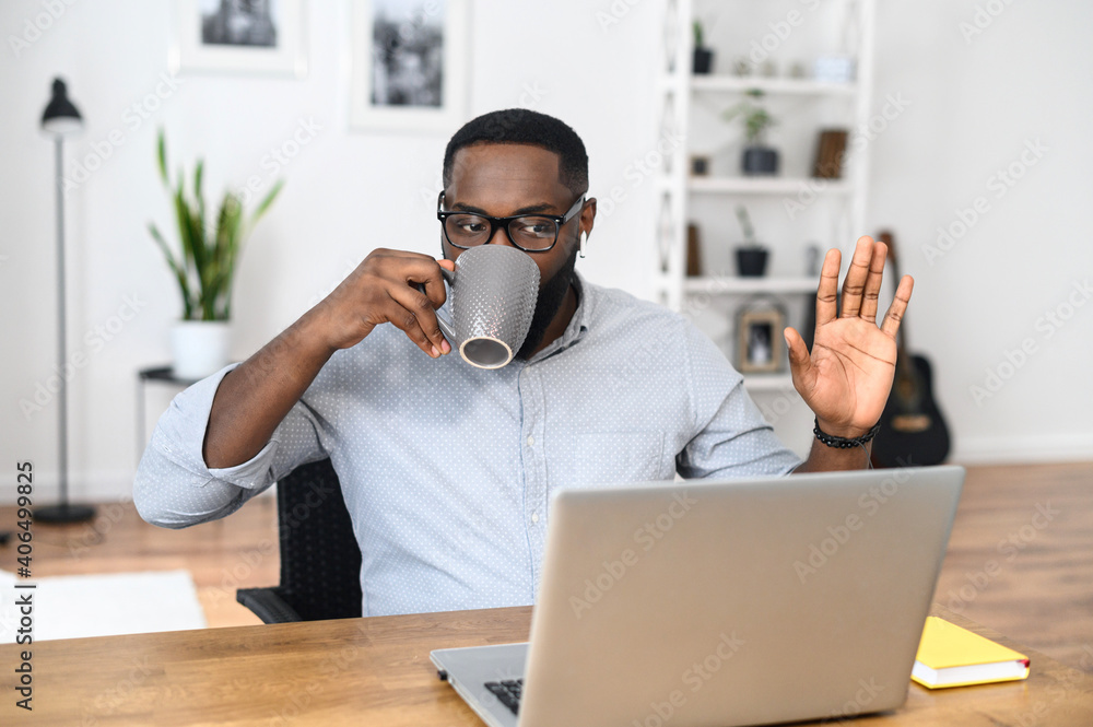 Young African-American businessman in glasses working remotely, sitting at the desk and drinking coffee, greeting participants with high-five, connected to the online conference via laptop Young African-American businessman in glasses working remotely, sitting at the desk and drinking coffee, greeting participants with high-five, connected to the online conference via laptop