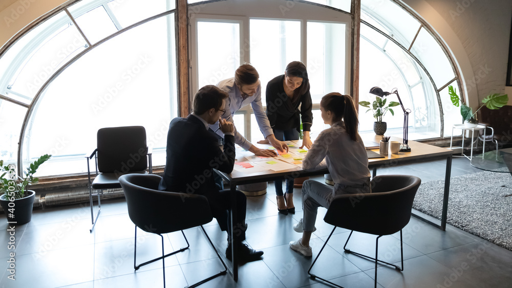 Diverse employees working with project documents, using sticky papers, writing notes, colleagues developing business project strategy, marketing plan, analyzing charts and diagrams in modern office