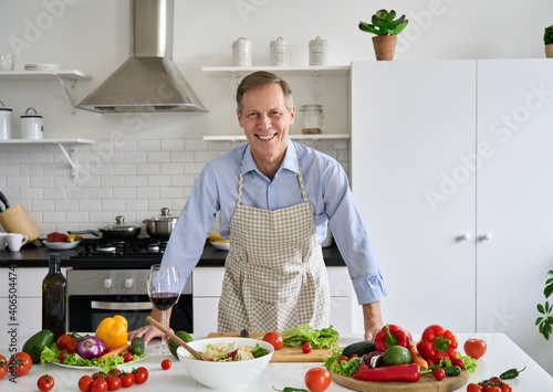 Smiling happy senior mature older 50s man wearing apron standing in kitchen table, looking at camera, preparing healthy food, making fresh vegetable salad enjoying cooking alone at home, portrait.