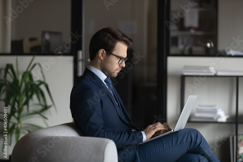 Side view confident businessman wearing glasses and suit working on laptop, sitting on couch in office, serious focused executive entrepreneur writing email, searching information, browsing apps