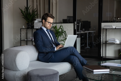 Confident businessman wearing suit and glasses working on laptop, sitting on couch in modern office, focused entrepreneur executive looking at computer screen, searching information, writing email
