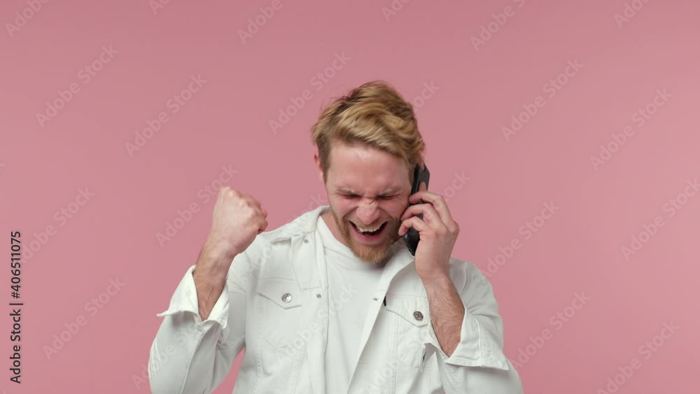 Excited happy bearded young man 20s years old in white casual clothes isolated on pastel pink background studio. People lifestyle concept. Talking on mobile phone doing winner gesture clenching fists Excited happy bearded young man 20s years old in white casual clothes isolated on pastel pink background studio. People lifestyle concept. Talking on mobile phone doing winner gesture clenching fists