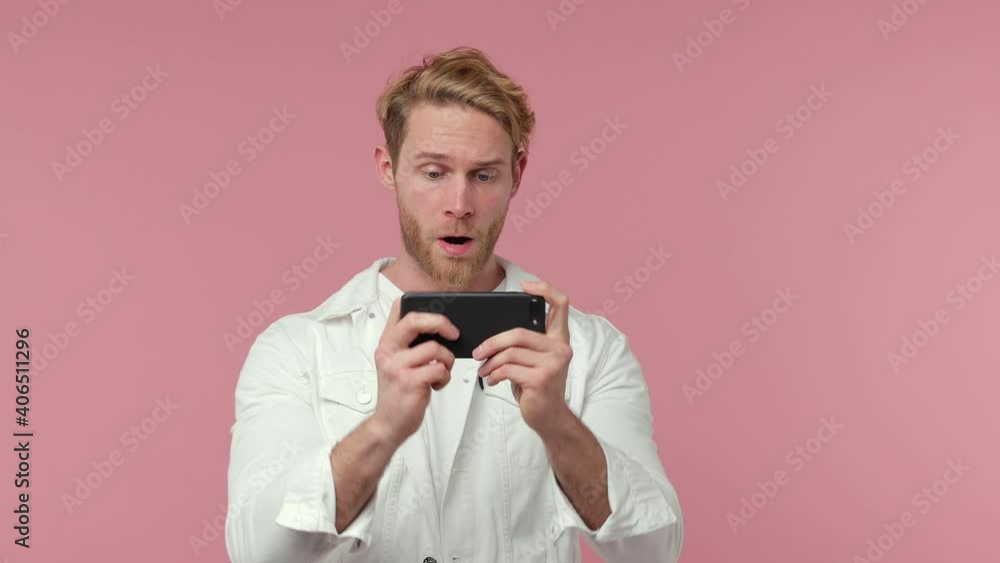 Excited cheerful bearded young man 20s years old in white casual clothes isolated on pink background studio. People lifestyle concept. Play game on mobile phone doing winner gesture clenching fists Excited cheerful bearded young man 20s years old in white casual clothes isolated on pink background studio. People lifestyle concept. Play game on mobile phone doing winner gesture clenching fists