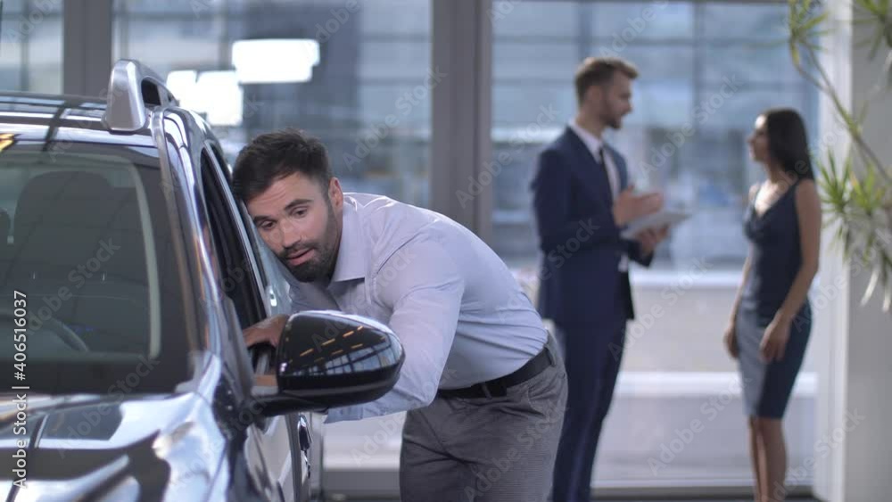 Handsome bearded man admiring car inside chosen for purchase in automobile showroom while his wife talking with auto dealer on background. Married couple choosing auto in car dealership Handsome bearded man admiring car inside chosen for purchase in automobile showroom while his wife talking with auto dealer on background. Married couple choosing auto in car dealership