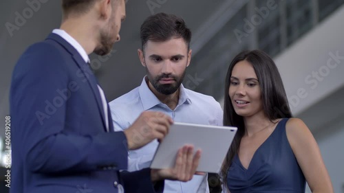 Close-up of professional salesman using tablet while showing couple of customers specifications of chosen car. Interested woman and man communicating with auto dealer and making choice at car showroom