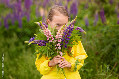 A girl in a yellow raincoat holds a bouquet of wild purple flowers. Flowers cover the girl's face. Field in the background