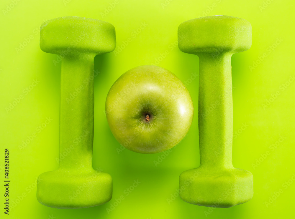 two green dumbbells and one green apple in the center, green background, the concept of sports and recuperation, keeping fit. two green dumbbells and one green apple in the center, green background, the concept of sports and recuperation, keeping fit.