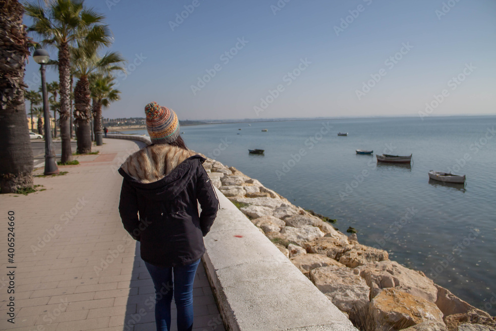 Beautiful girl with mask and hat next to the beach in winter with boats in the water

