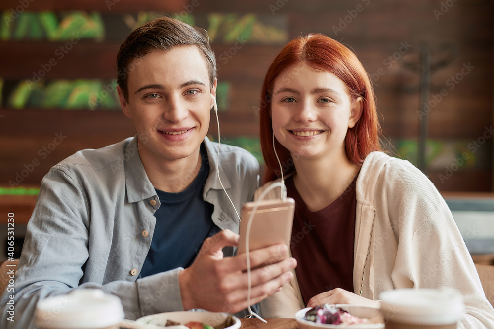 Portrait of teenage couple smiling at camera while listening to music together using smartphone and same pair of earphones, sitting in a cafe on a daytime