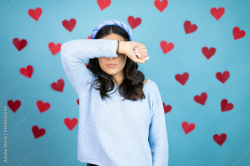 Young beautiful woman over blue background with red hearts covering eyes with arm smiling cheerful and funny. Blind concept. Young beautiful woman over blue background with red hearts covering eyes with arm smiling cheerful and funny. Blind concept.