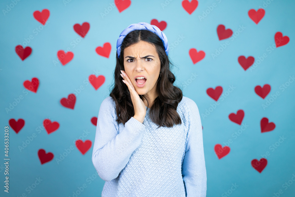 Young beautiful woman over blue background with red hearts touching mouth with hand with painful expression because of toothache or dental illness on teeth Young beautiful woman over blue background with red hearts touching mouth with hand with painful expression because of toothache or dental illness on teeth