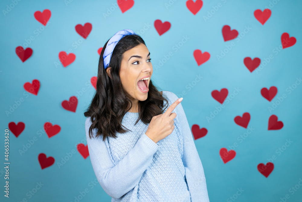 Young beautiful woman over blue background with red hearts amazed and pointing with hand and finger to the side Young beautiful woman over blue background with red hearts amazed and pointing with hand and finger to the side