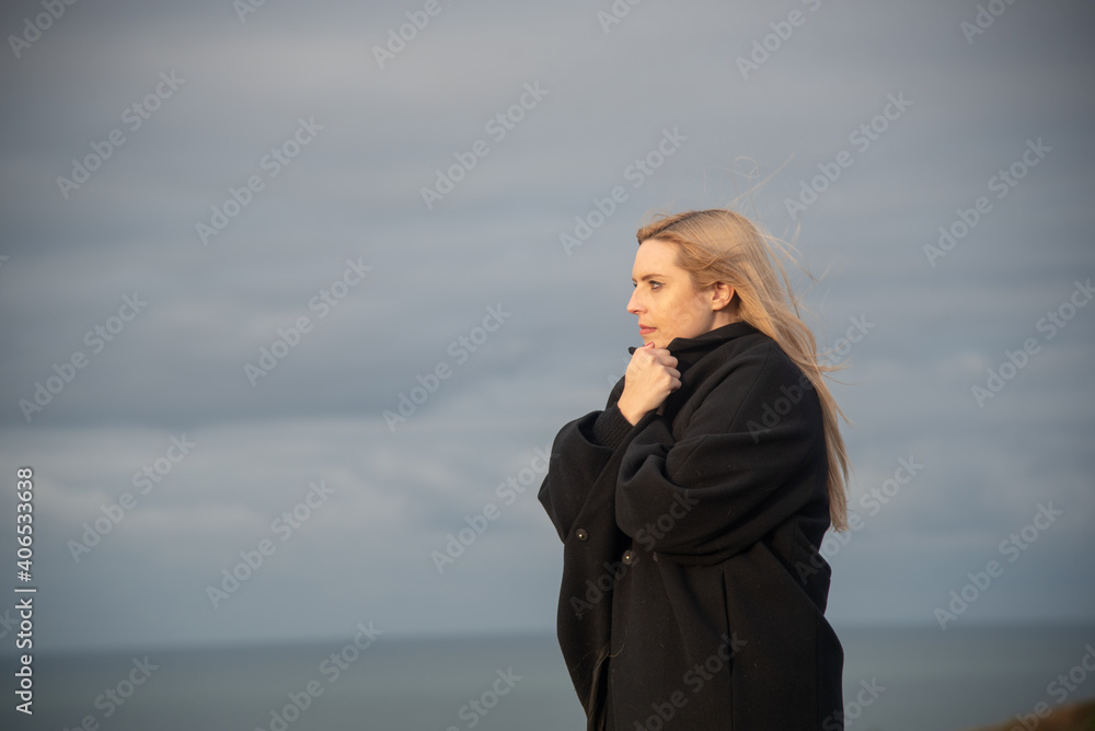 mujer rubia con abrigo negro mirando hacia el mar y tomando el aire en un día de invierno y cielo nublado mujer rubia con abrigo negro mirando hacia el mar y tomando el aire en un día de invierno y cielo nublado