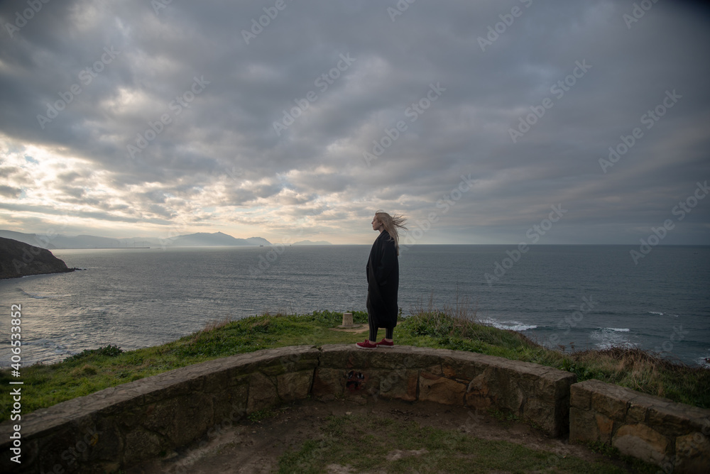 mujer rubia con abrigo negro mirando hacia el mar y tomando el aire en un día de invierno y cielo nublado