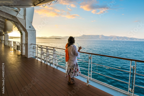 A woman sips a drink on the deck of a cruise ship as the sun sets and the ship passes islands on the Aegean Sea