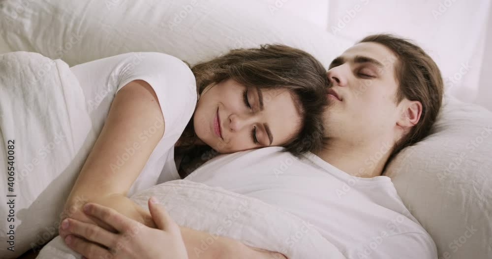 Young Caucasian couple enjoying good quality sleep, resting together in bed. Loving woman wife sweetly cuddling her husband, smiling. Young Caucasian couple enjoying good quality sleep, resting together in bed. Loving woman wife sweetly cuddling her husband, smiling.