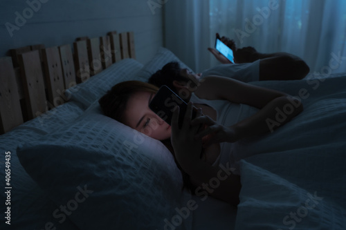 Asian woman and Caucasian man lay on bed use smartphone for social media and online at night. Copy space concept of diversity couple, relationship and communication problem, social media addiction.