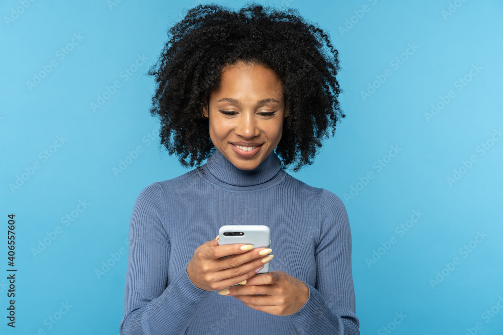 Stylish happy African millennial woman with curly hair wear blue turtleneck, reading message from boyfriend, chatting in social media, texting sms, isolated on studio blue background. Stylish happy African millennial woman with curly hair wear blue turtleneck, reading message from boyfriend, chatting in social media, texting sms, isolated on studio blue background.