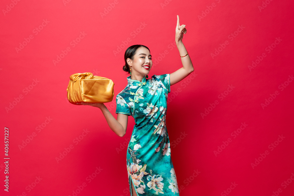 Pretty Asian woman in oriental style costume holding gold gift box pointing hand upward in red isolated studio background for Chinese new year concepts Pretty Asian woman in oriental style costume holding gold gift box pointing hand upward in red isolated studio background for Chinese new year concepts