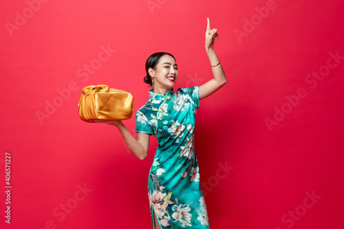 Pretty Asian woman in oriental style costume holding gold gift box pointing hand upward in red isolated studio background for Chinese new year concepts
