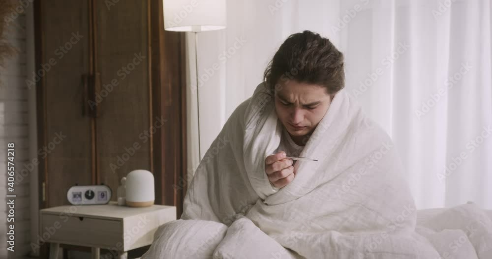 Sick young man staying in bed, wrapped in warm blanket, suffering from fever, mesuring temperature. Coronavirus pandemic. Sick young man staying in bed, wrapped in warm blanket, suffering from fever, mesuring temperature. Coronavirus pandemic.