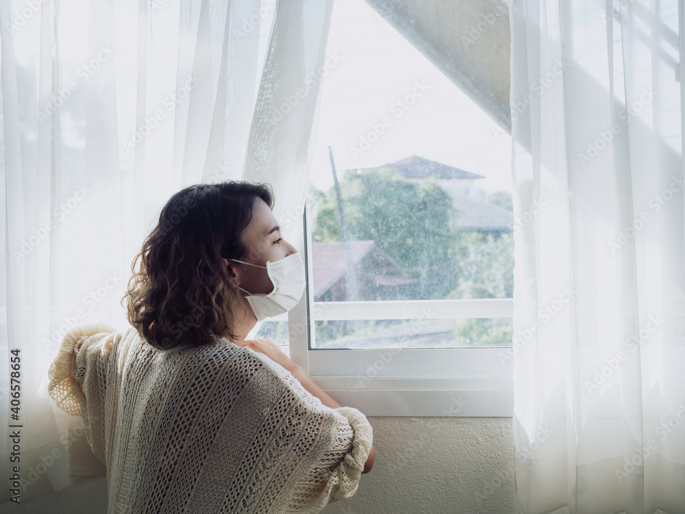 Beautiful lonely Asian woman wearing medical face mask looking out the window.