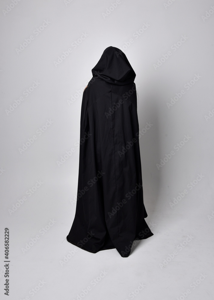 Full length portrait of pretty black haired woman wearing long dark gown nada cloak. Standing pose facing away from the camera, against a studio background. Full length portrait of pretty black haired woman wearing long dark gown nada cloak. Standing pose facing away from the camera, against a studio background.