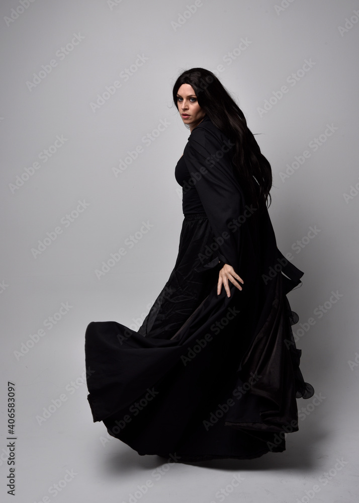 Full length portrait of pretty black haired woman wearing long dark gown nada cloak. Standing pose facing away from the camera, against a studio background. Full length portrait of pretty black haired woman wearing long dark gown nada cloak. Standing pose facing away from the camera, against a studio background.