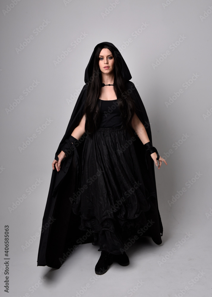 Full length portrait of pretty black haired woman wearing long dark gown and a cloak.  Standing pose facing away from the camera, against a  studio background.