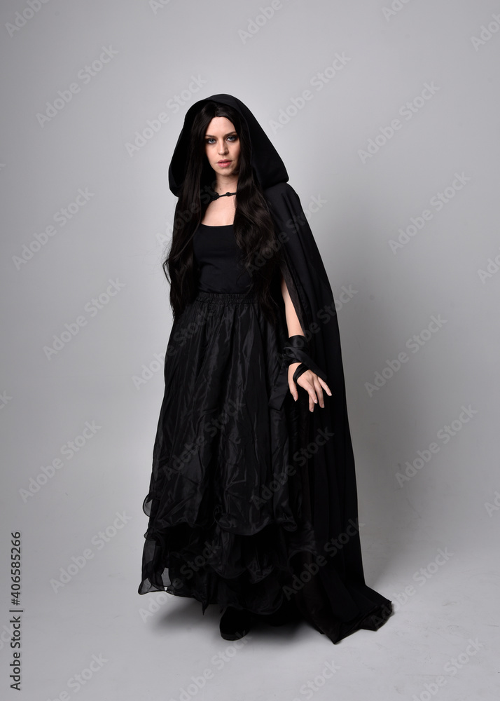 Full length portrait of pretty black haired woman wearing long dark gown and a cloak. Standing pose facing away from the camera, against a studio background. Full length portrait of pretty black haired woman wearing long dark gown and a cloak. Standing pose facing away from the camera, against a studio background.