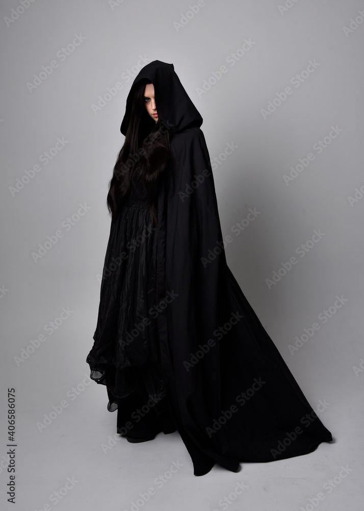 Full length portrait of pretty black haired woman wearing long dark gown and a cloak. Standing pose facing away from the camera, against a studio background. Full length portrait of pretty black haired woman wearing long dark gown and a cloak. Standing pose facing away from the camera, against a studio background.