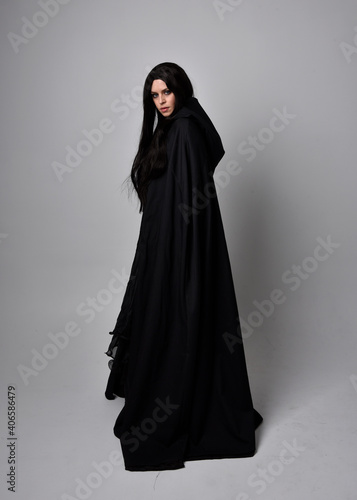 Full length portrait of pretty black haired woman wearing long dark gown and a cloak. Standing pose facing away from the camera, against a studio background.